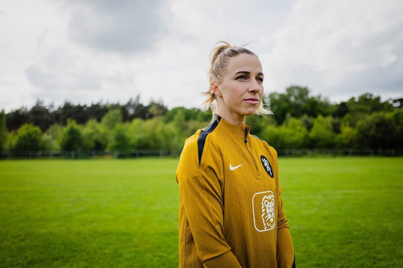 Woman in gold Nike sports top on green field, looking right.