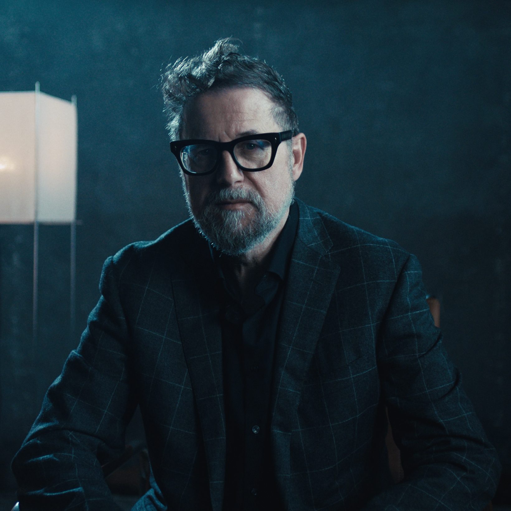 A man with a beard and glasses, wearing a dark plaid suit jacket, looks directly at the viewer.