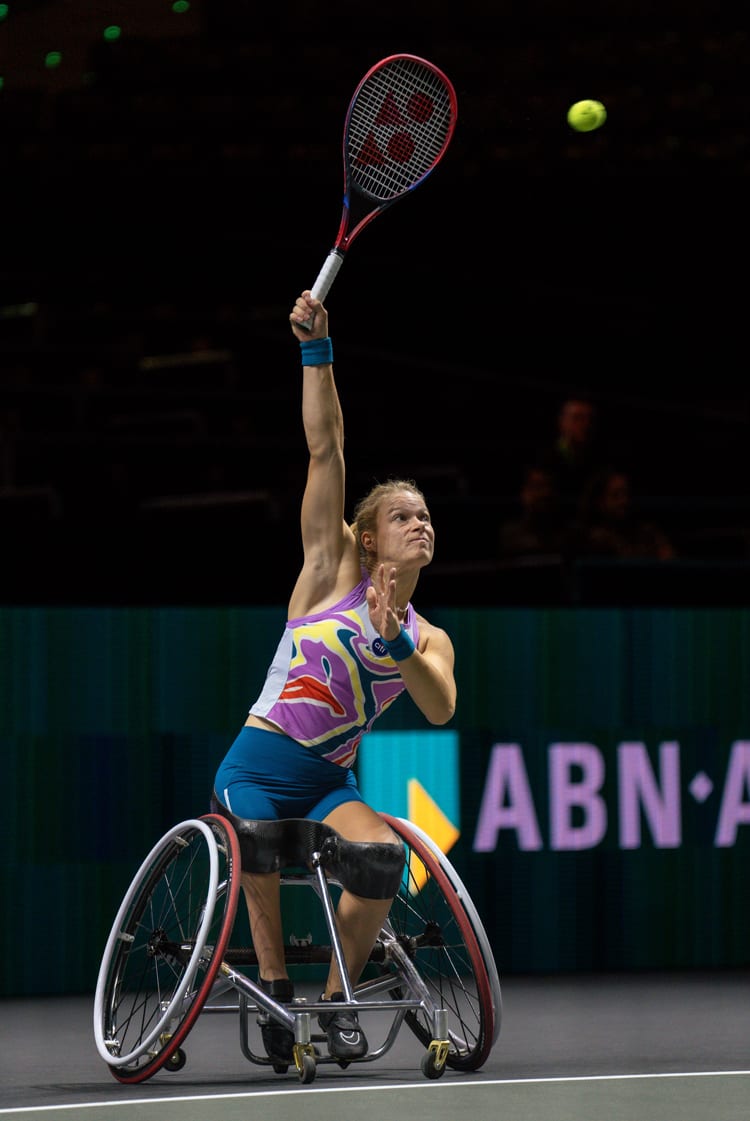 A female wheelchair tennis player in a colorful top raises her racket to serve, eyes focused on the tennis ball.