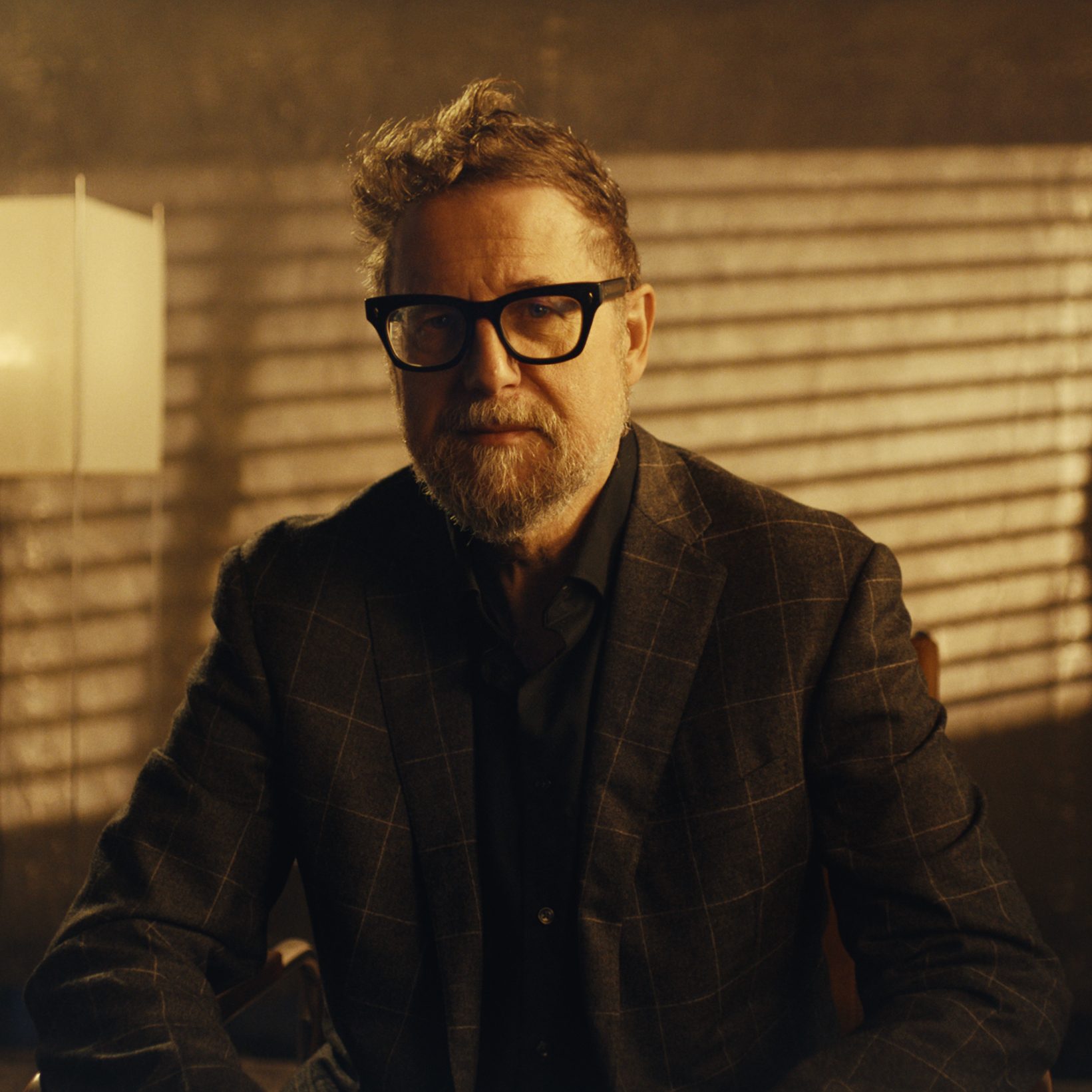 A man with a beard and glasses in a plaid suit looks directly at the camera in a warmly lit room.