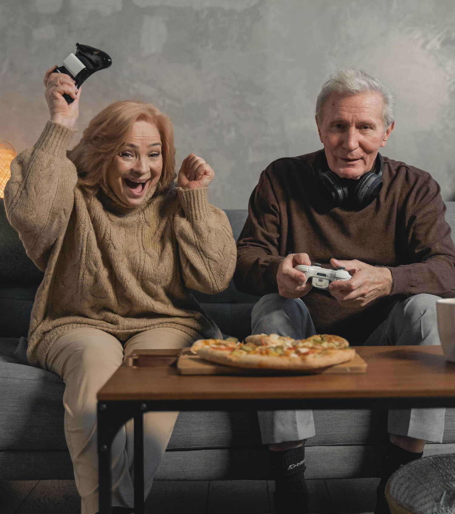 Two seniors joyfully game on a couch with pizza; she's cheering, he's concentrating.