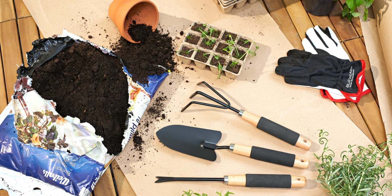 Gardening supplies including soil, tools, seedlings, and gloves on a wooden and paper surface.