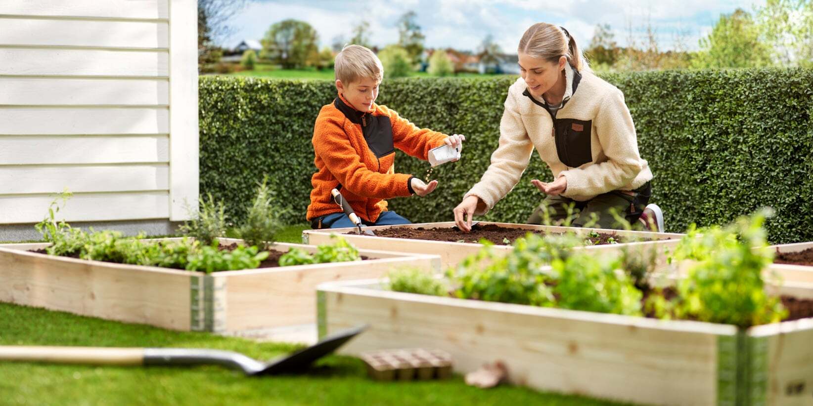 A woman and a boy planting seeds in wooden raised garden beds on a sunny day.