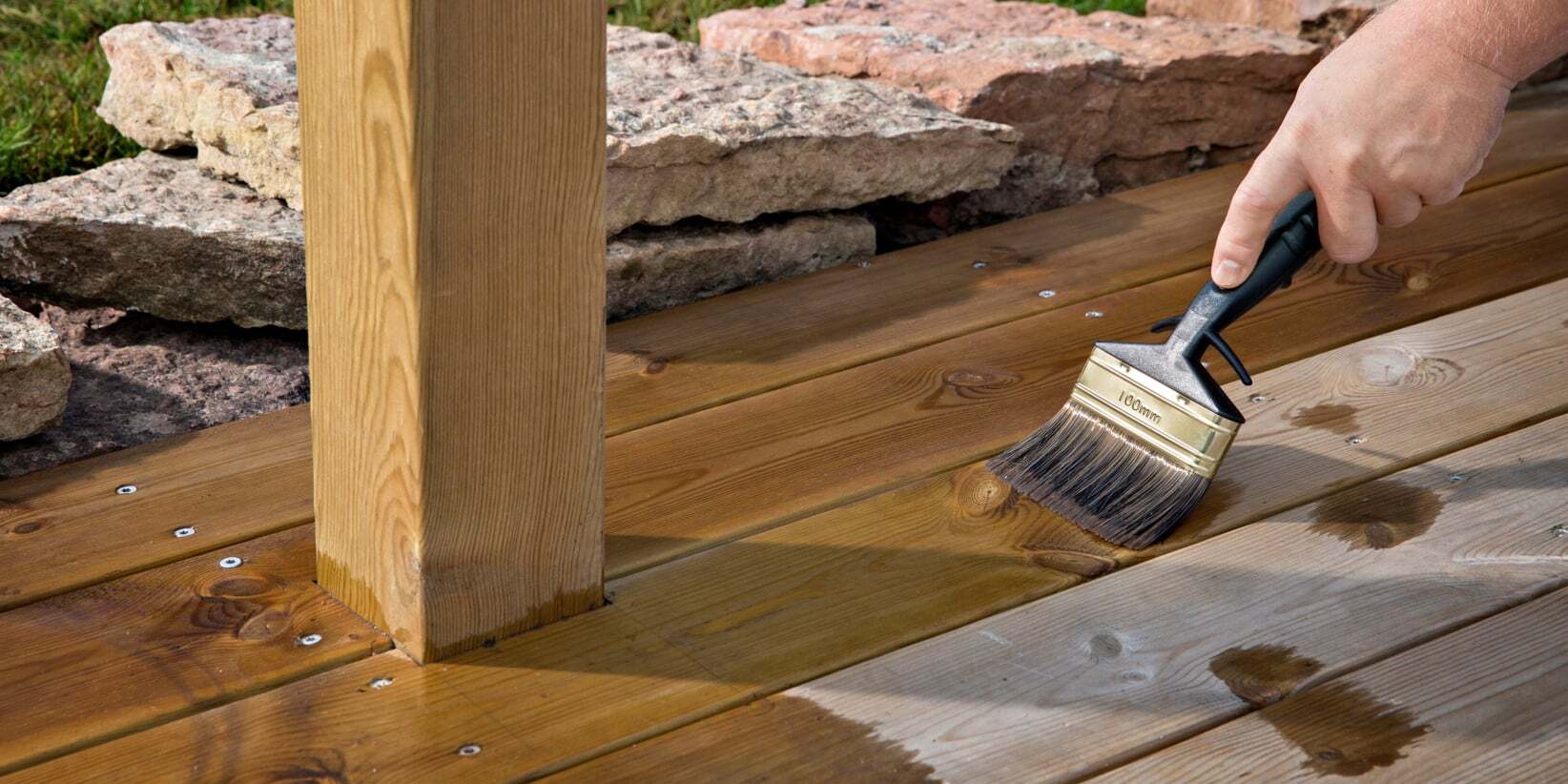 A hand staining a wooden deck with a brush, showing the contrast between treated and untreated wood.