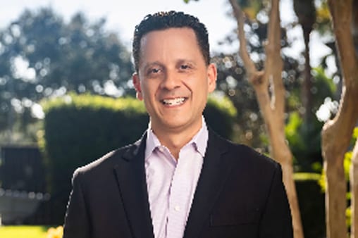 Smiling man in a dark suit jacket and light purple shirt, standing outdoors among green foliage.