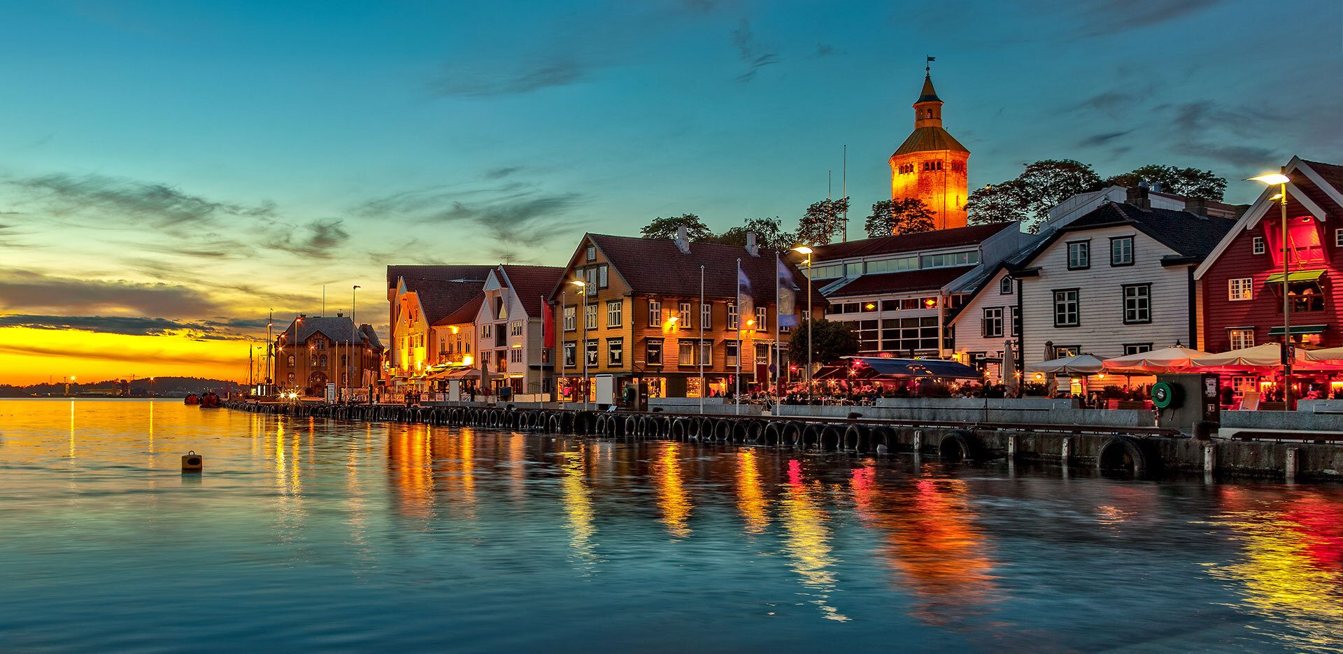 Waterfront town at dusk with illuminated buildings, water reflections, and a tower.