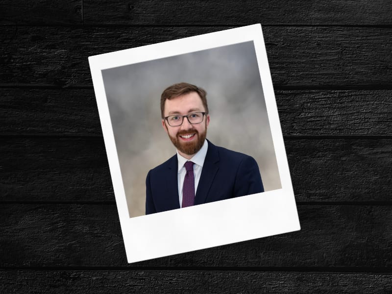 Headshot of a smiling man with glasses and a beard, wearing a suit and patterned tie, on a gray background.