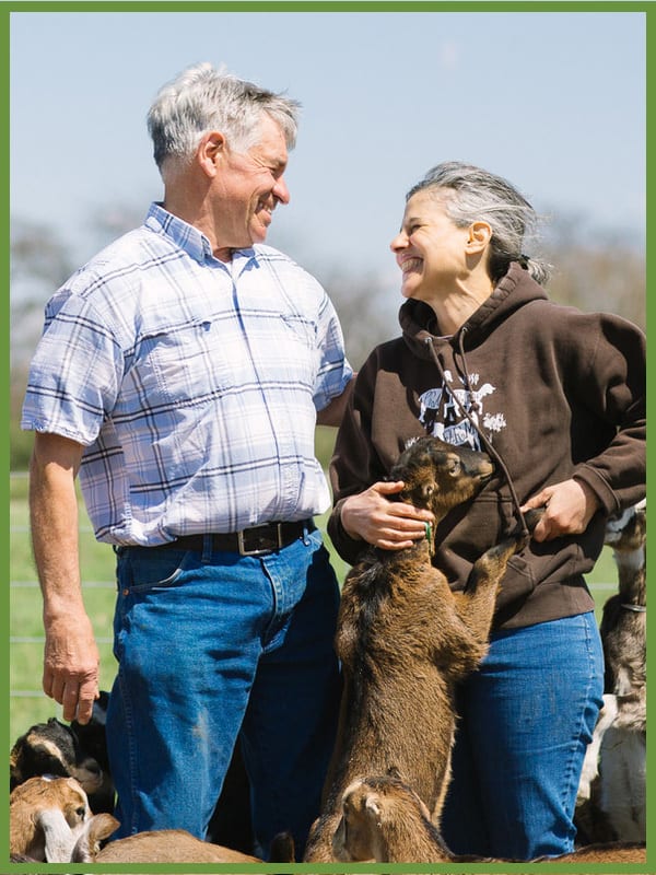 Smiling man and woman with multiple baby goats; one goat stands with the woman.