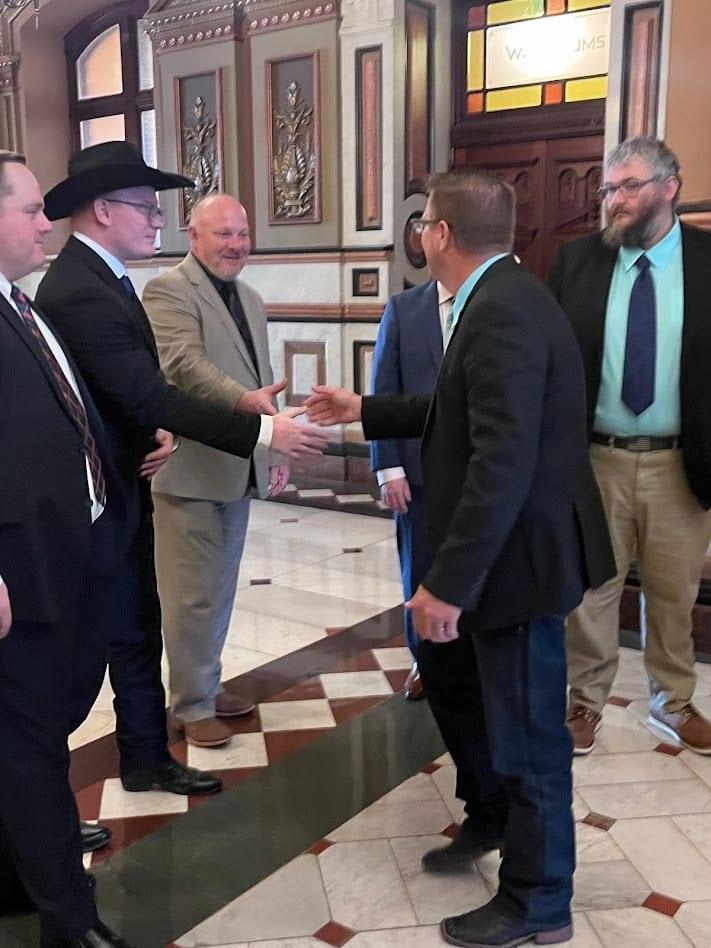 Several men in suits, including one in a cowboy hat, shake hands in a grand hallway.