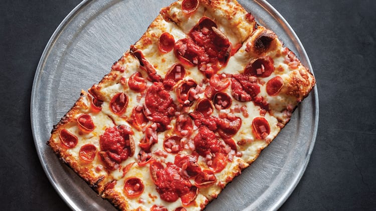 Rectangular pizza with pepperoni, cheese, and diced ham on a round metal serving tray.