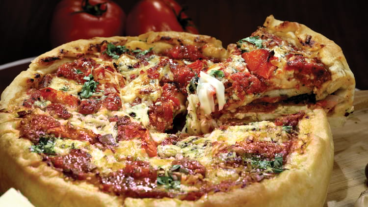 Deep-dish pizza, slice removed with cheese pull, rich tomato sauce, and herbs.
