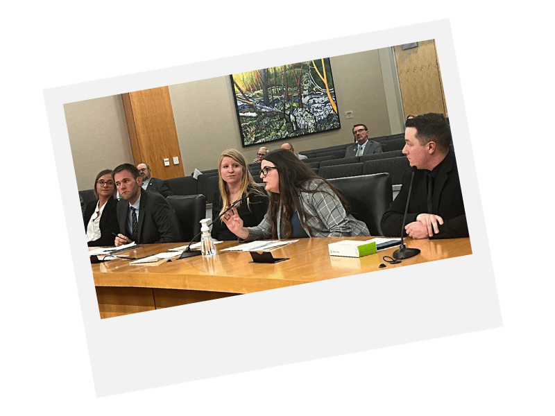People at a conference table with microphones; a woman in a plaid jacket is speaking.
