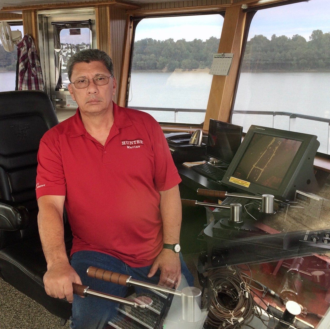 A man in a red polo shirt sits in the wheelhouse of a boat, overlooking a river.