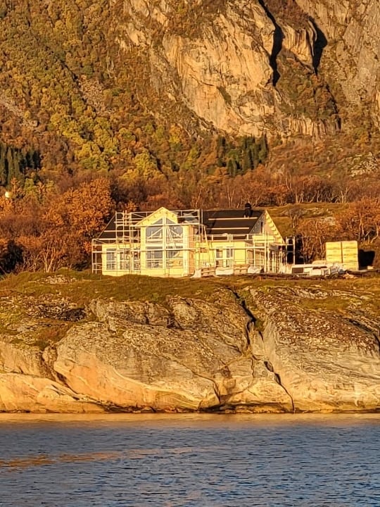 New house under construction on a rocky shore by water, backed by autumn mountains.
