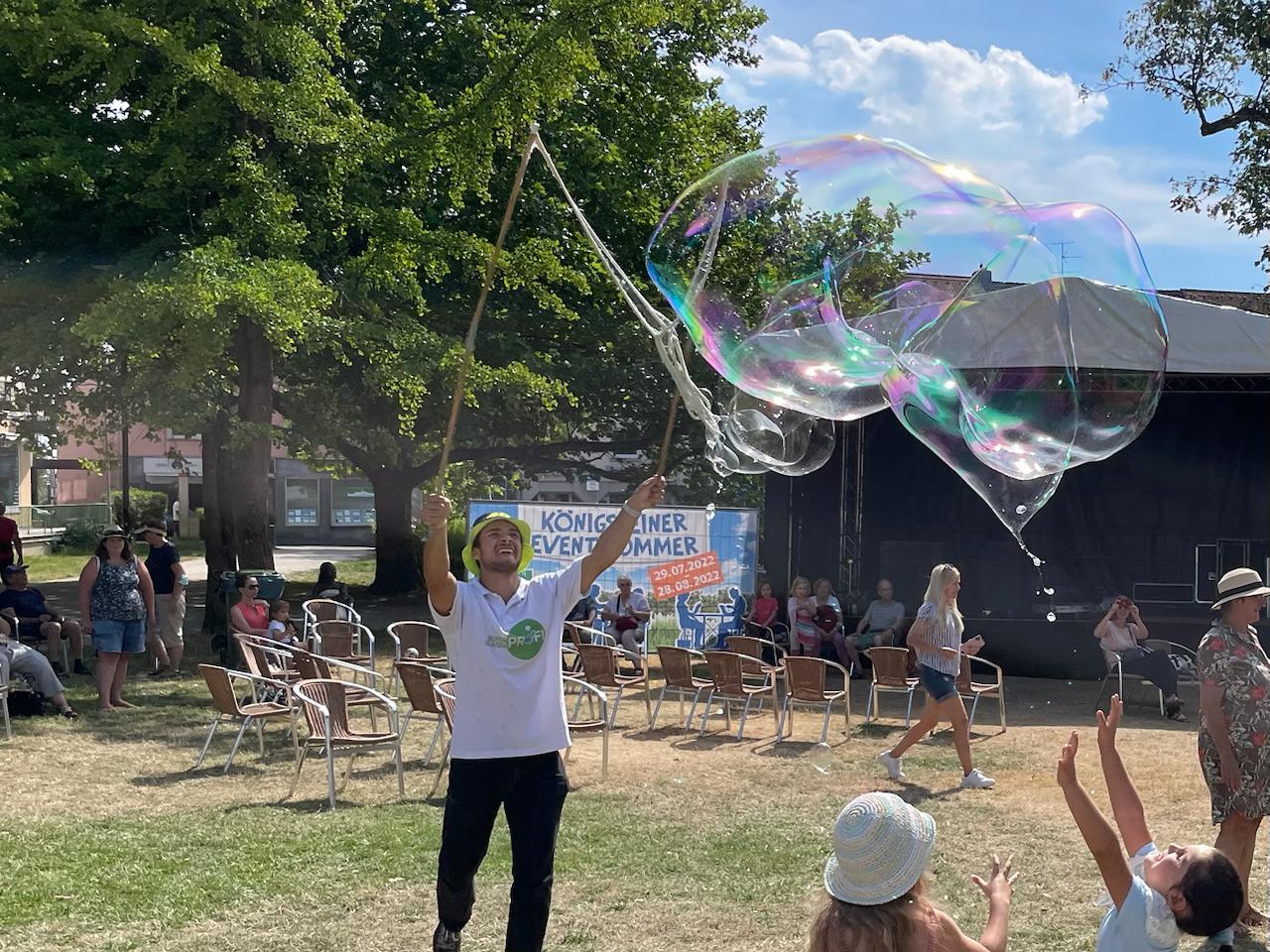 A man in a yellow hat and white t-shirt makes huge iridescent bubbles at an outdoor summer event, with children eagerly reaching for them.
