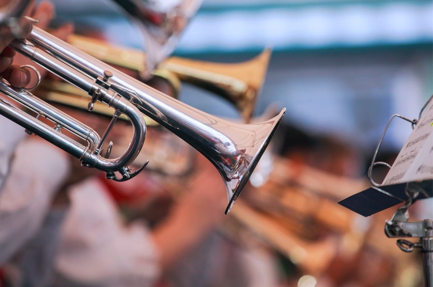 Silver trumpet played by a musician, with blurred band and sheet music in the background.