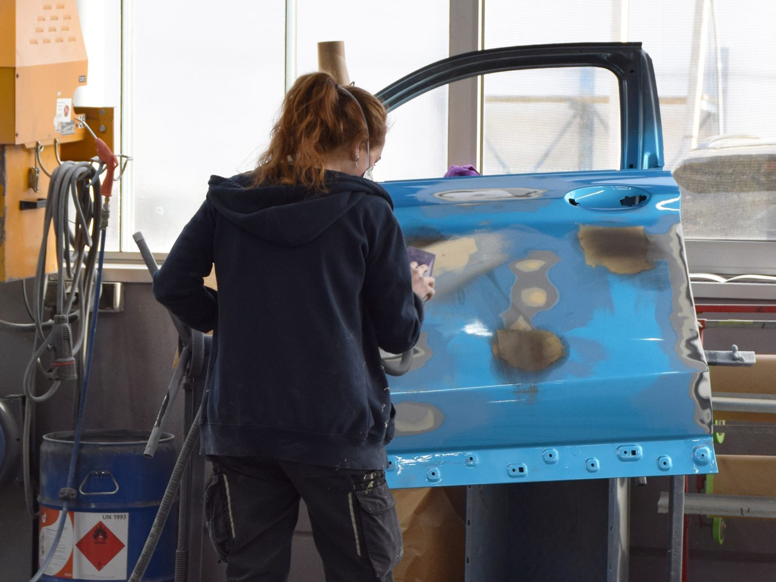 A person sands a blue car door in a body shop, prepping it for paint.