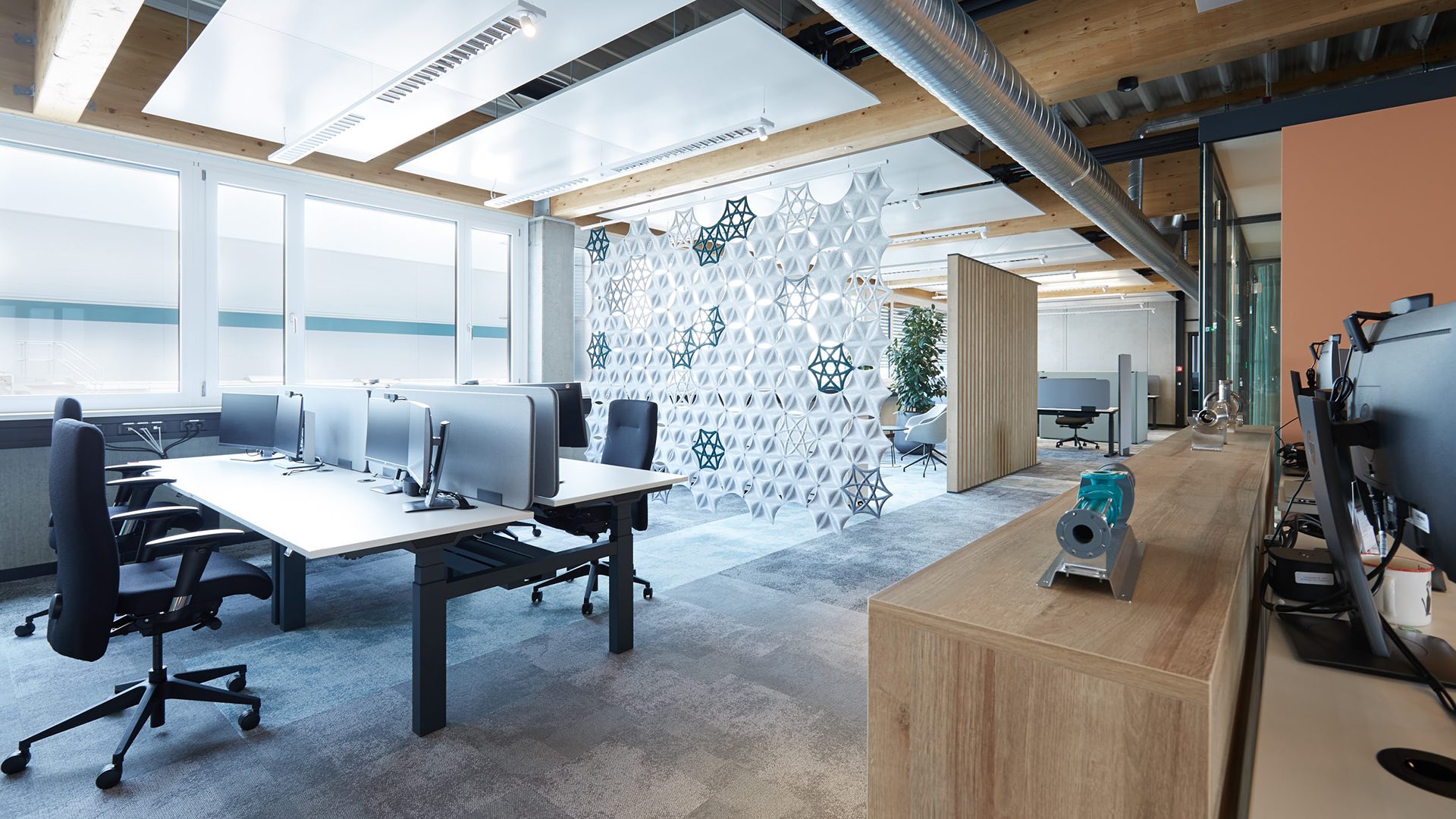 Modern open-plan office with computers, chairs, large windows, and a decorative geometric partition.