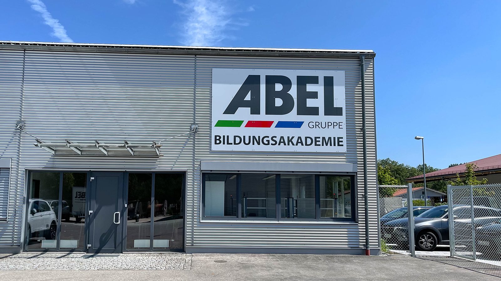 Building with "ABEL GRUPPE BILDUNGSAKADEMIE" sign, glass entrance, and parked cars.