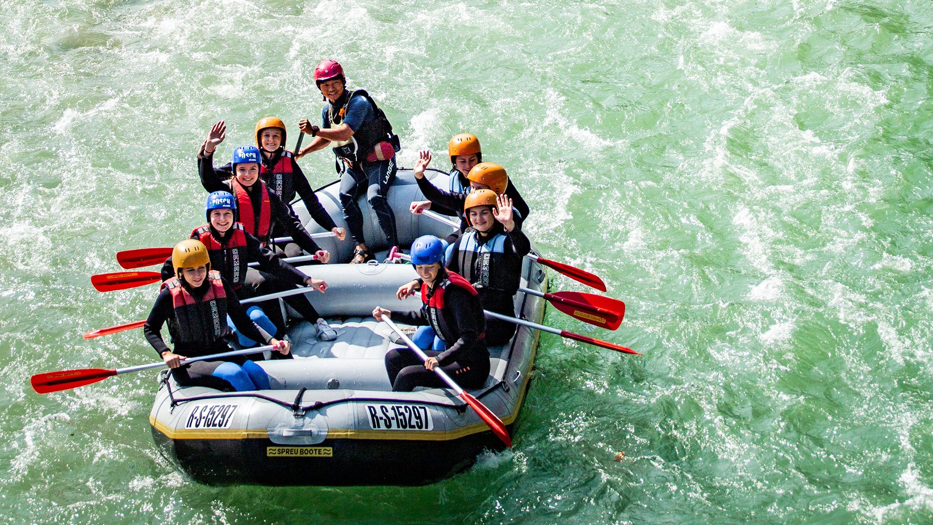 Seven people in helmets and life vests wave from a raft on a rushing green river.