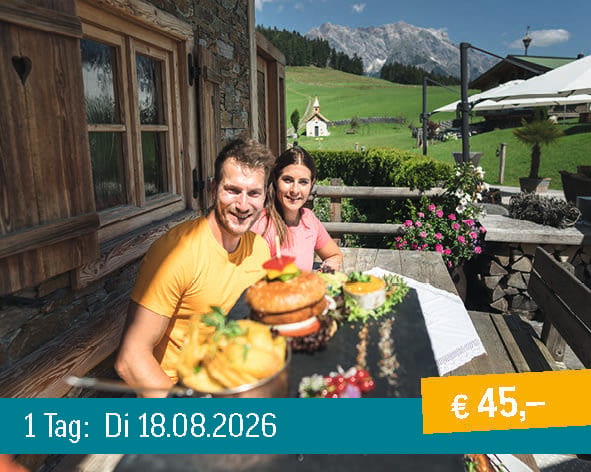 A smiling couple enjoys a meal with a burger and fries on a mountain terrace with a scenic view.