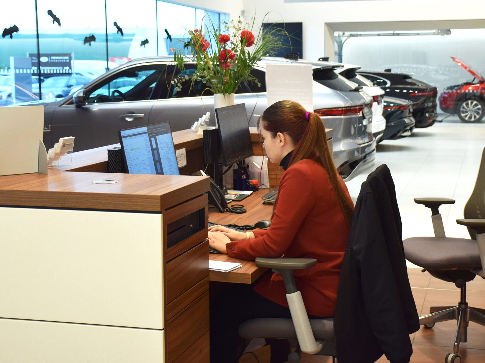 A woman in a red jacket works at a desk with computers in a modern car showroom.