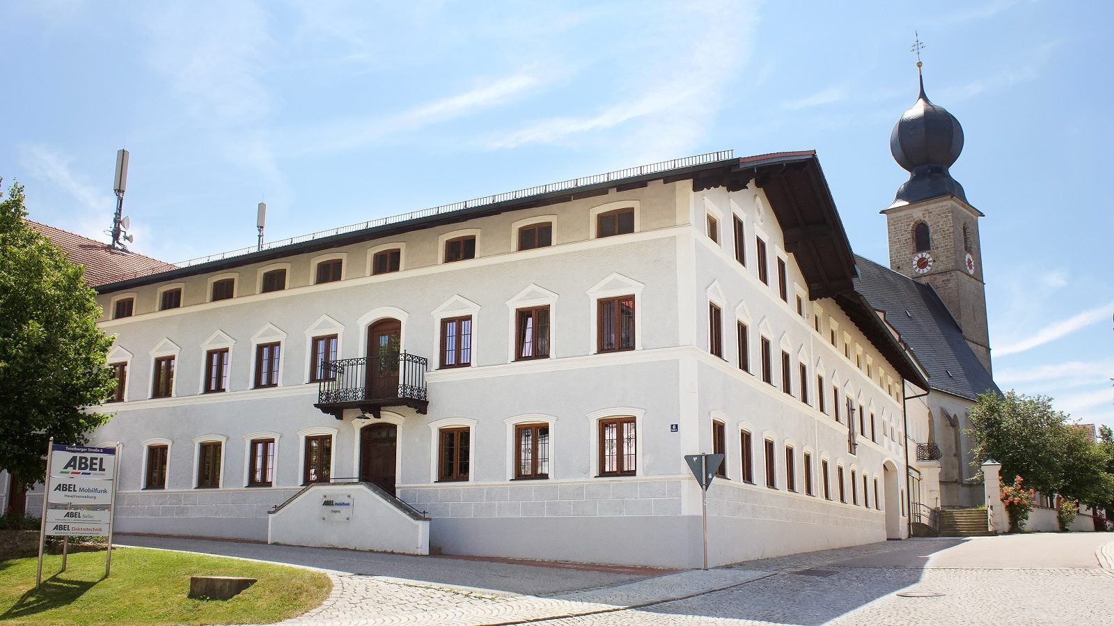 A large white building with brown windows and a nearby church tower under a sunny sky.
