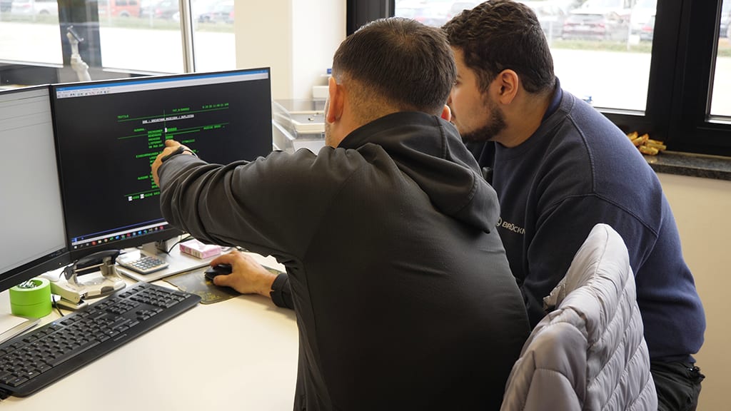Two men collaborate at a computer, with one pointing to the screen showing green text.