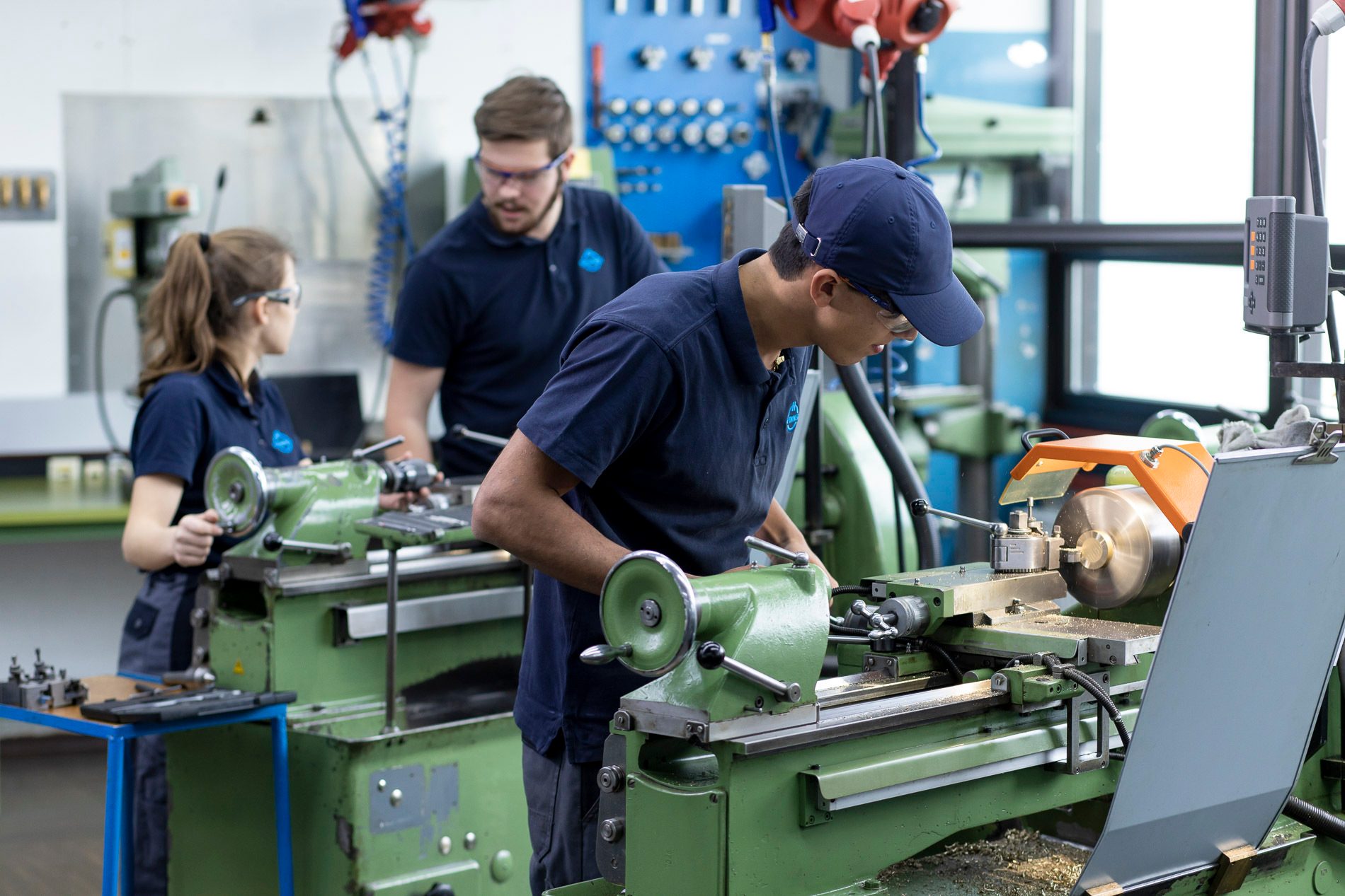Three people operating green lathes in a workshop, wearing safety glasses and dark blue uniforms.