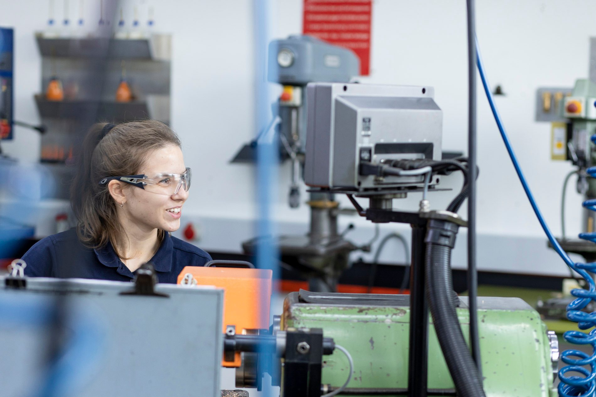 Smiling woman in safety glasses in a workshop with industrial machinery.