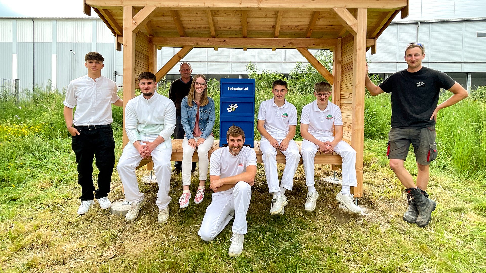 Group of smiling people with a wooden shelter and a blue box with a bee logo in a grassy field.