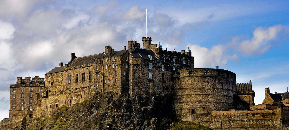 Edinburgh Castle, a historic fortress on a rocky crag, stands prominently against a cloudy blue sky.