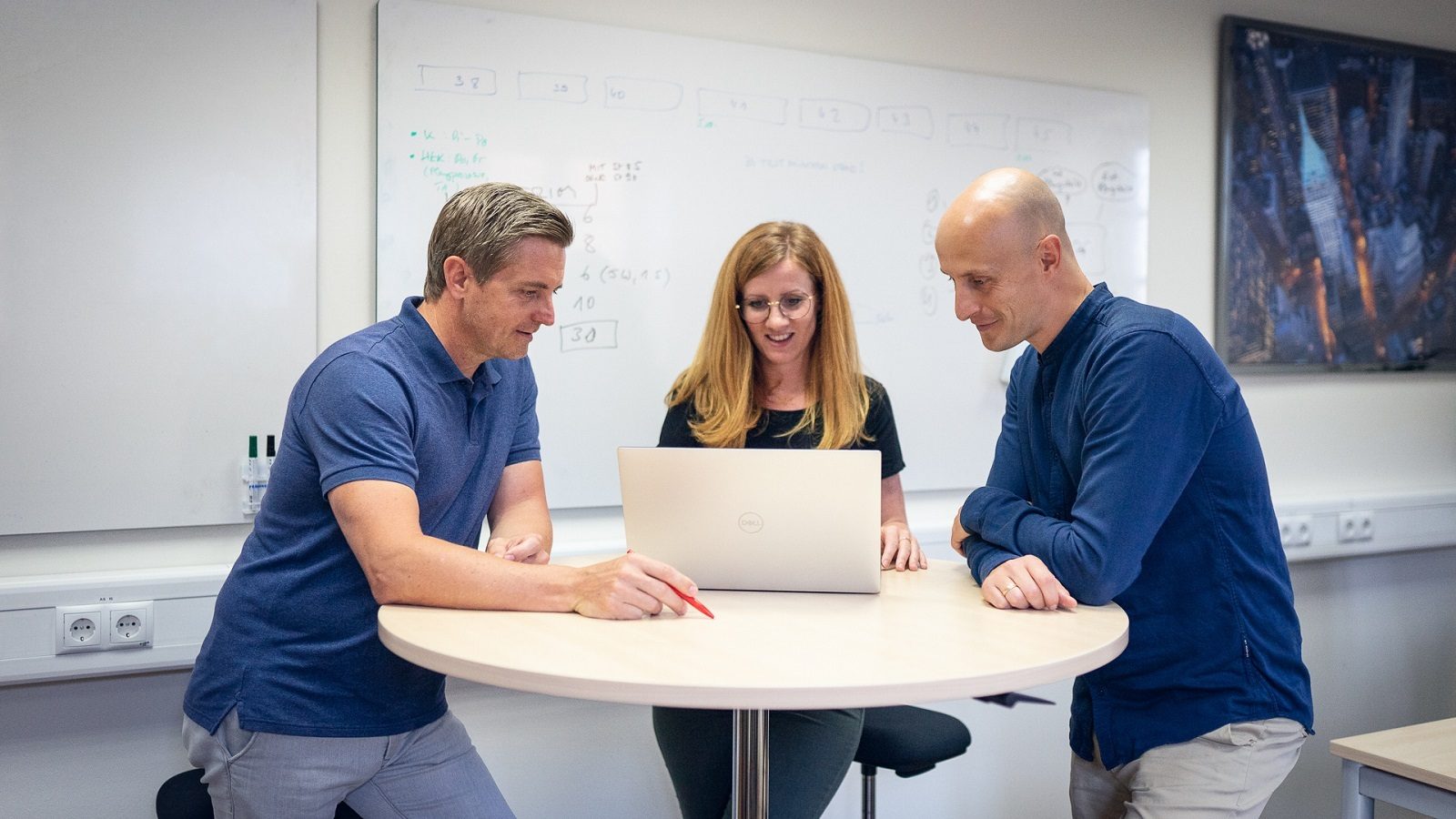 Three colleagues (two men, one woman) collaborate on a laptop at a high table in an office.