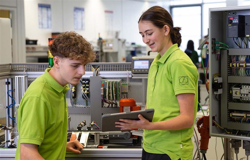 Two people in green shirts examine control systems with a tablet.