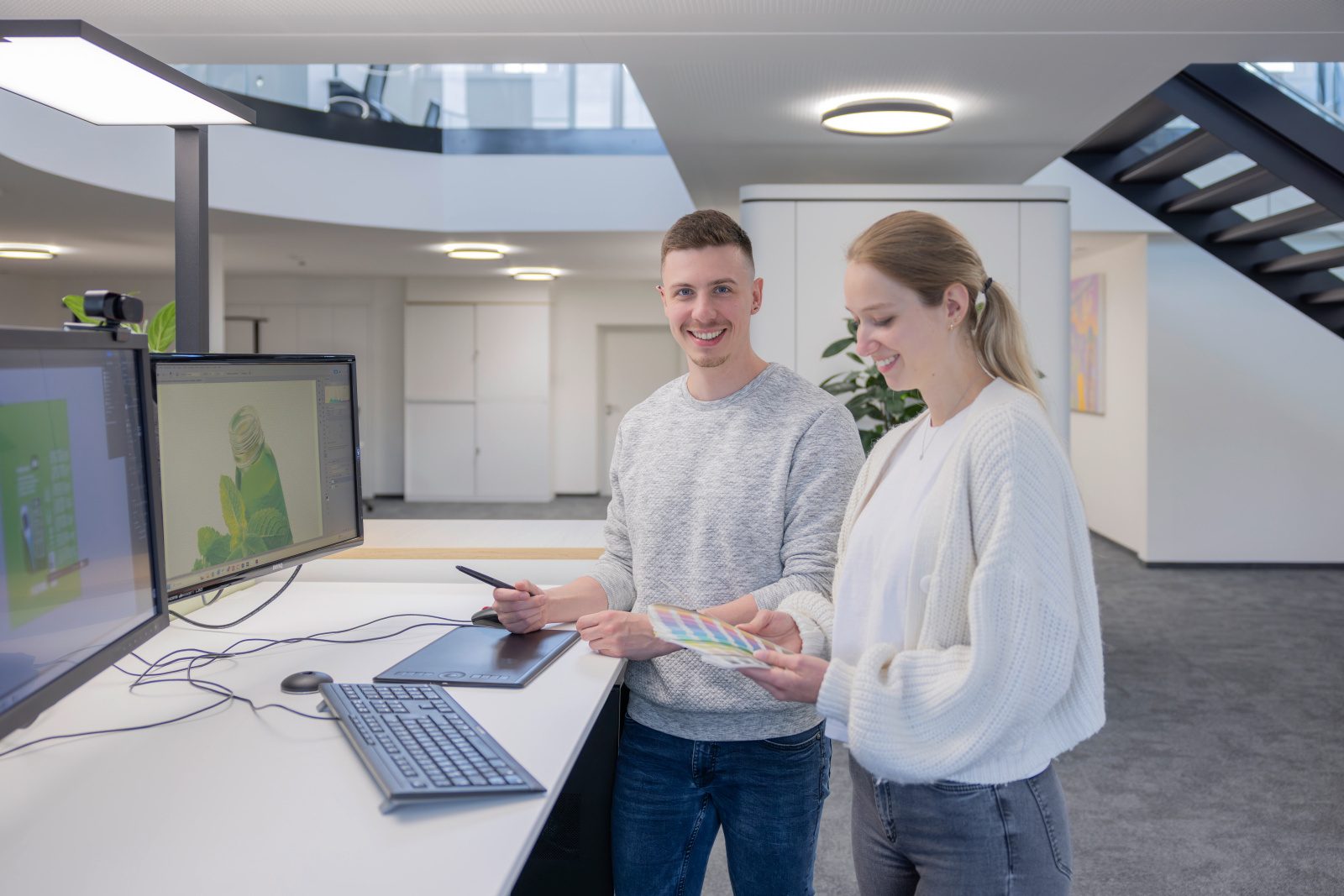 Two smiling designers collaborate at a desk with computers, a graphics tablet, and a color palette.