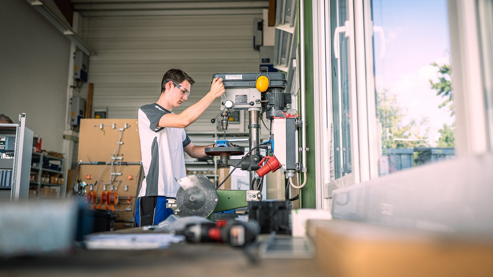 Young man in safety glasses drilling metal with a drill press in a workshop.