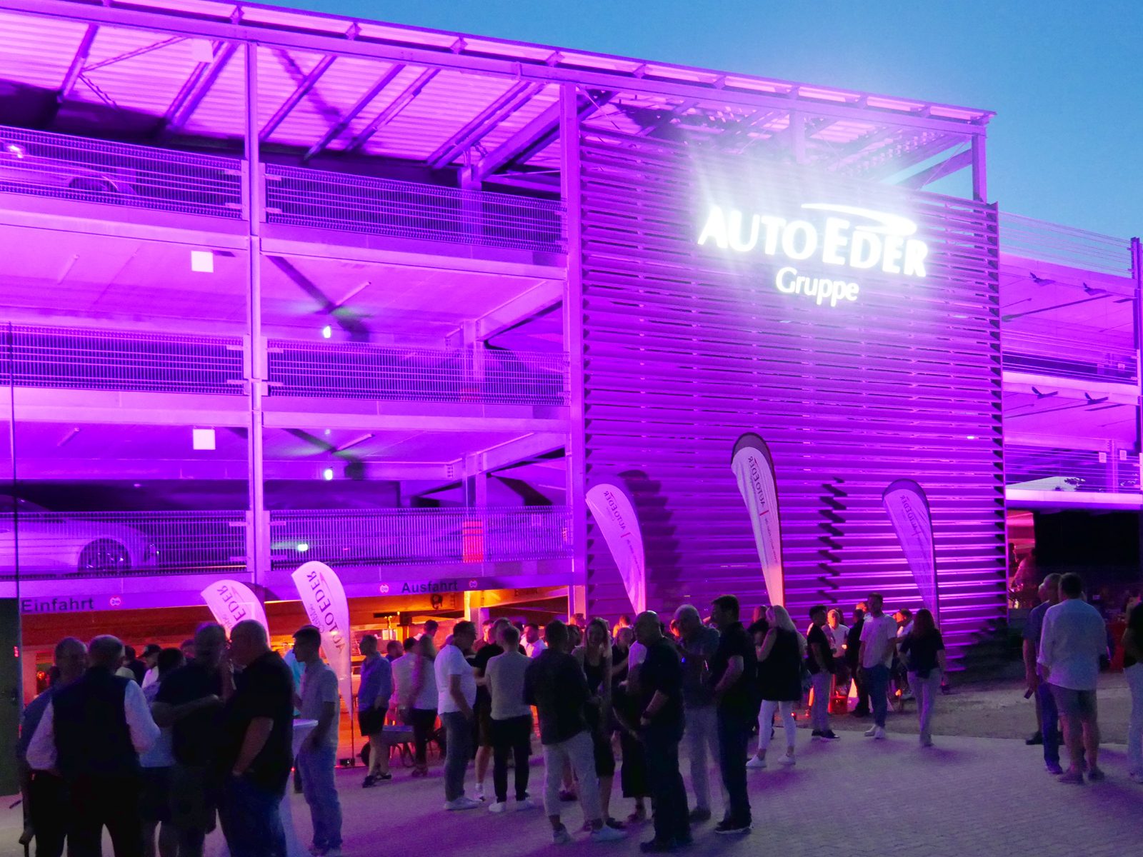Night view of a purple-lit building with "AUTO EDER Gruppe" sign, surrounded by a crowd.