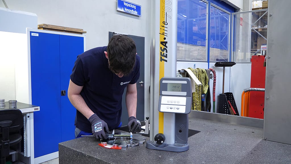 Man measuring a metal part with a caliper next to a TESA hite 700 measuring device in a workshop.