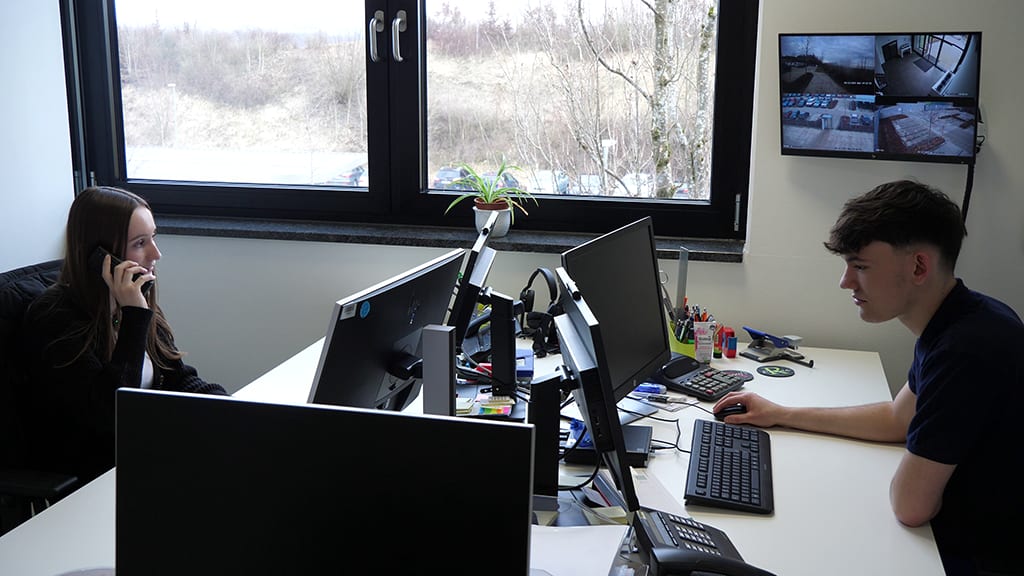 Two people at desks in an office: a woman on the phone, a man on a computer.