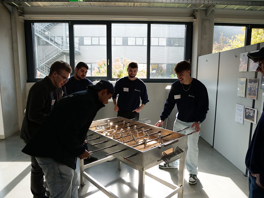 Young men playing foosball in a well-lit room with large windows.