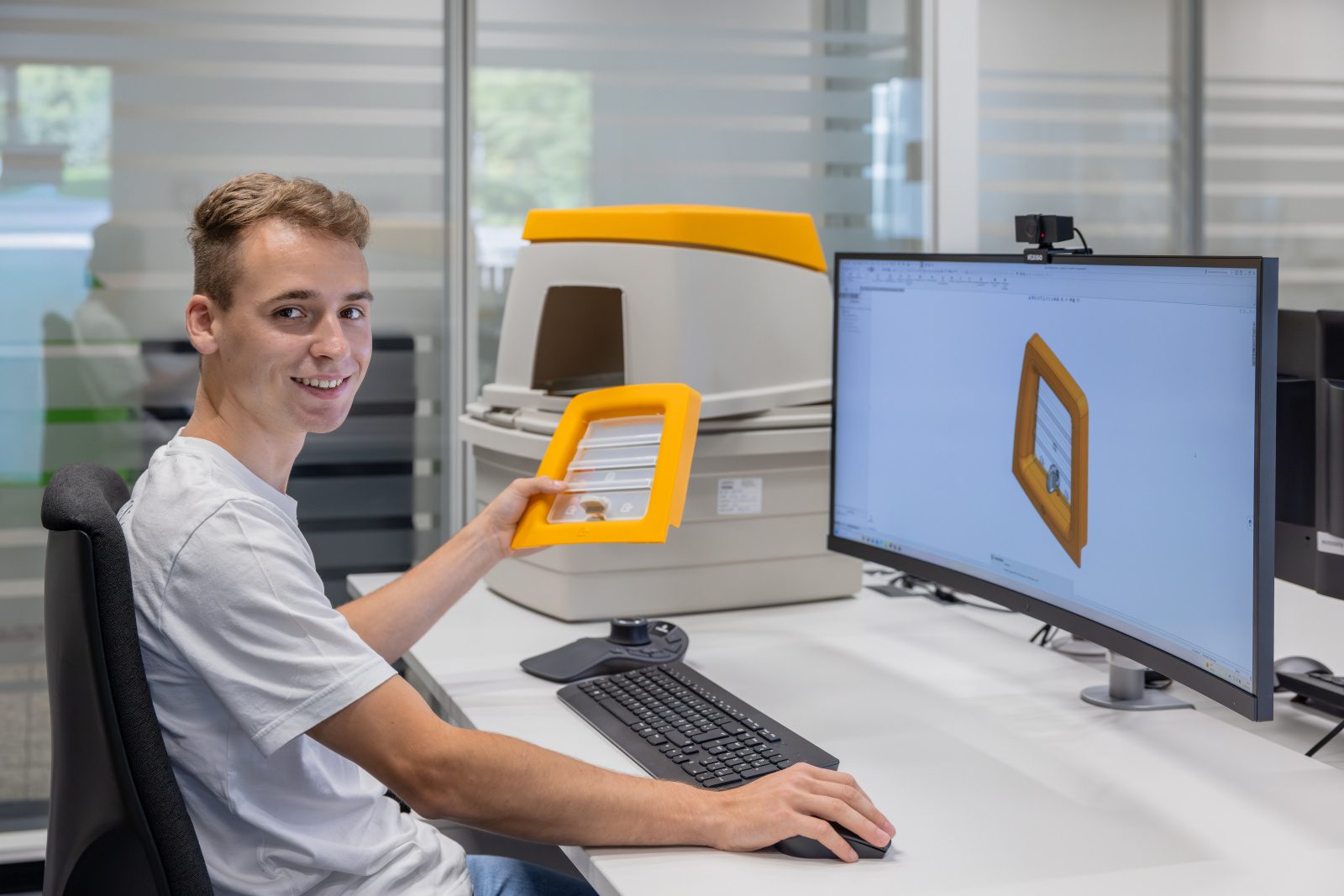 A young man smiles, holding an orange part, with its 3D model displayed on his computer screen.