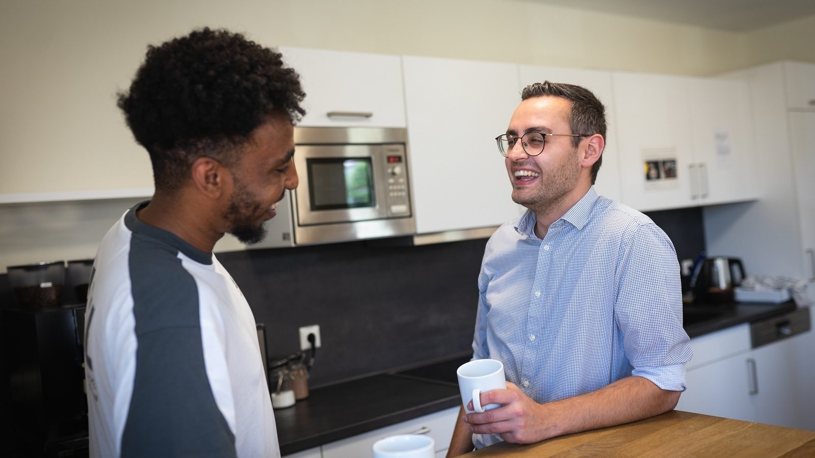 Two diverse men laughing in a kitchen, one holding a mug.