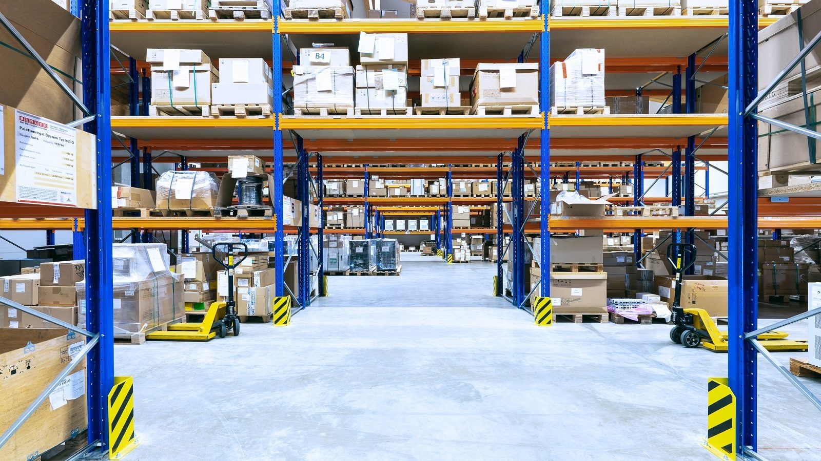 Bright warehouse with high blue/orange racks stacked with boxes and pallets, and yellow pallet jacks.