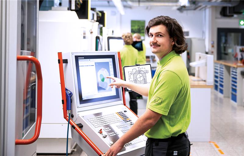 Man in green shirt points at CAD on CNC machine screen, holding tablet with technical drawing.