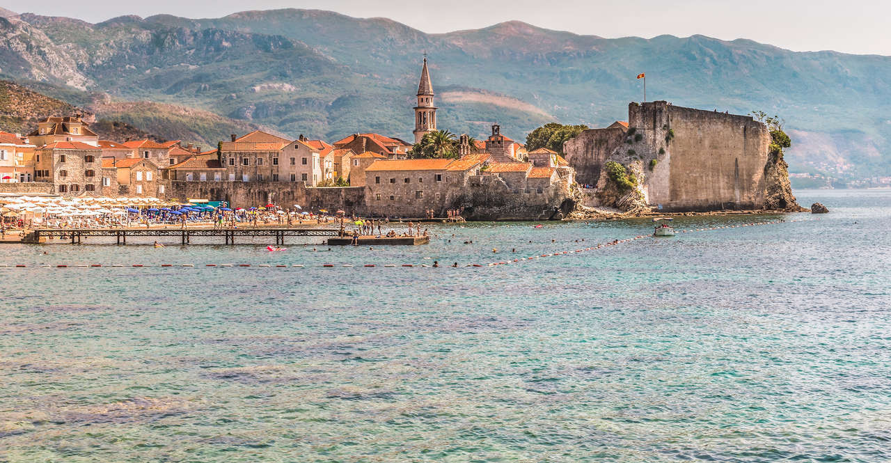 Historic coastal town with a prominent church tower, fortress, beach with umbrellas, and clear blue sea.