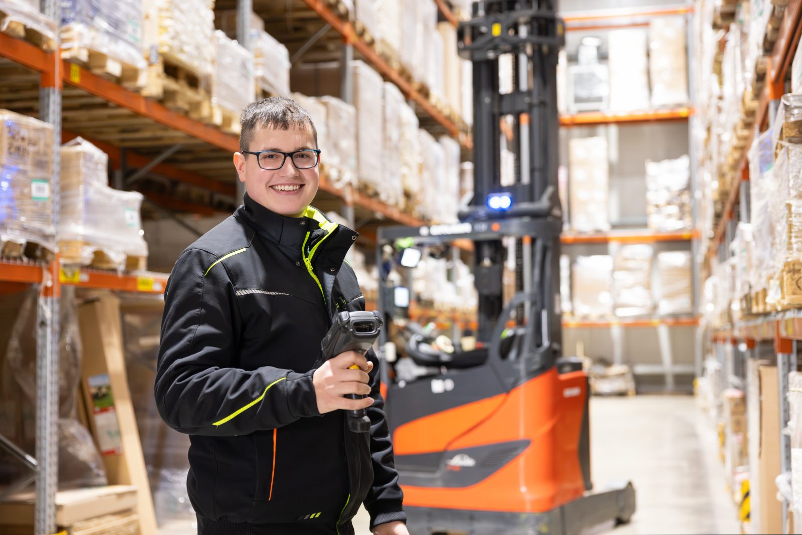Smiling man in a warehouse holding a scanner, with a forklift nearby.