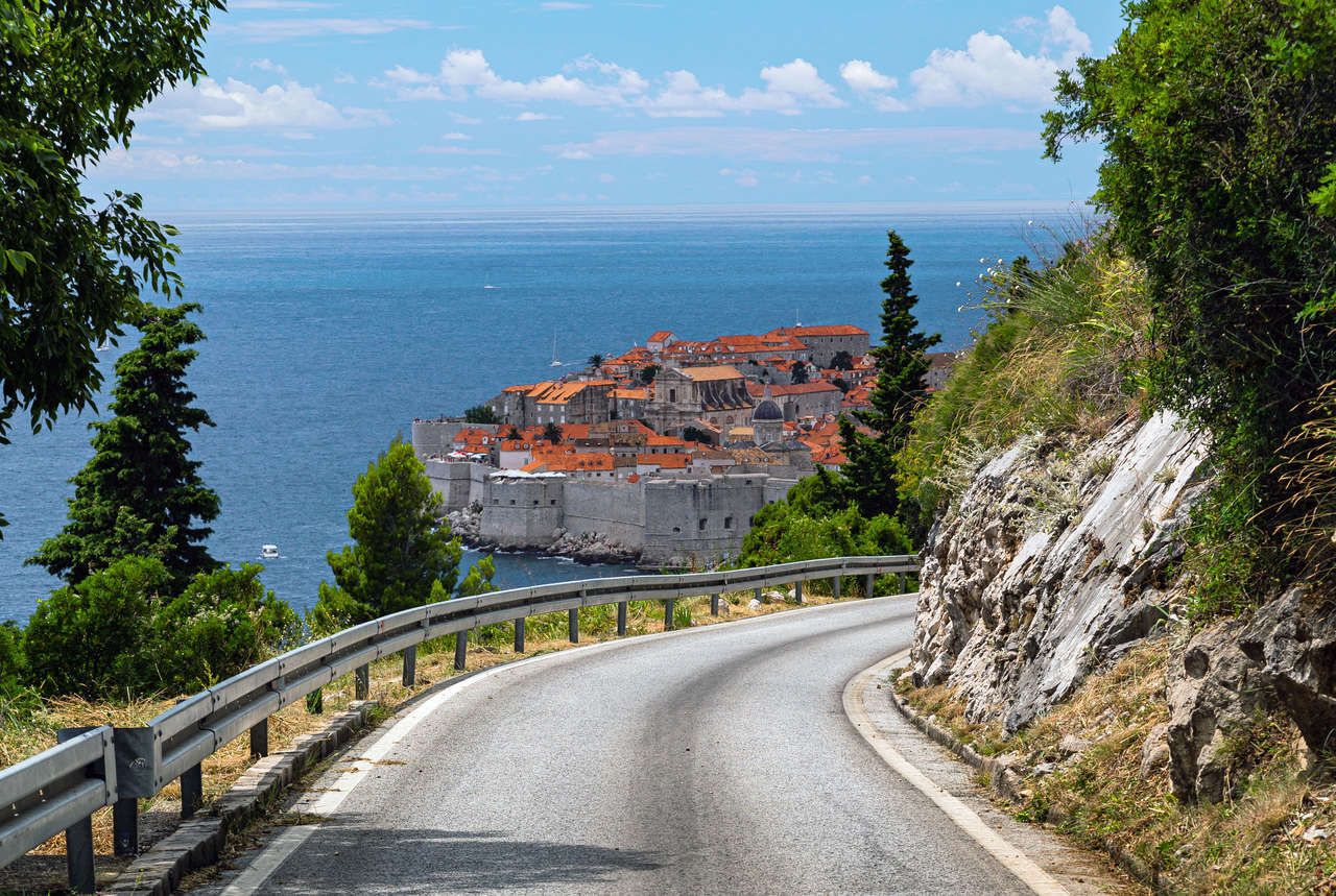A winding coastal road leads to Dubrovnik's historic walled city and the blue Adriatic Sea under a clear sky.