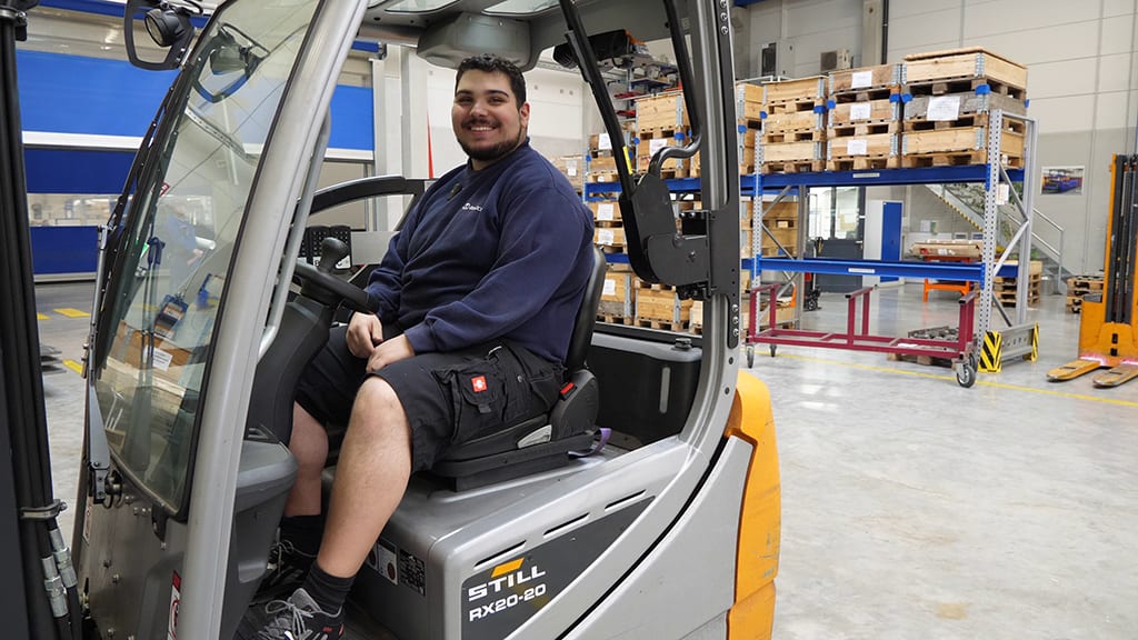 Smiling man sits in a STILL forklift in a warehouse with stacked pallets.