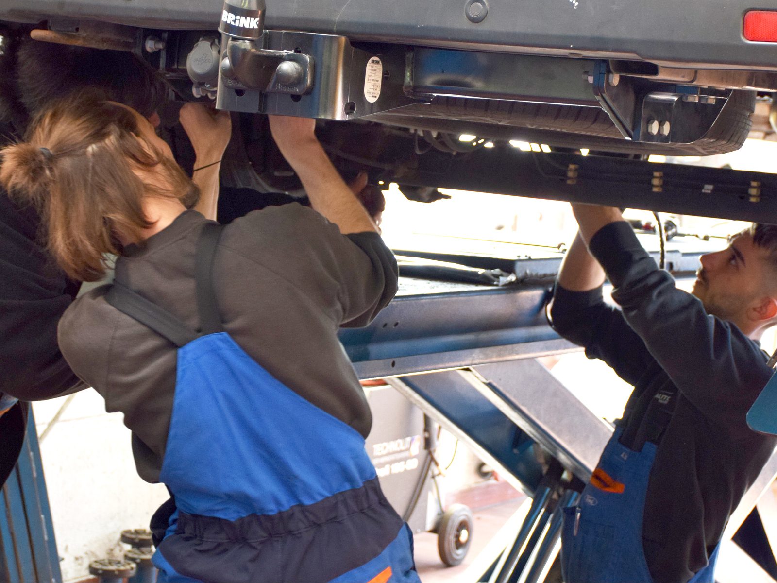 Two mechanics in overalls installing a Brink tow hitch on a lifted car.