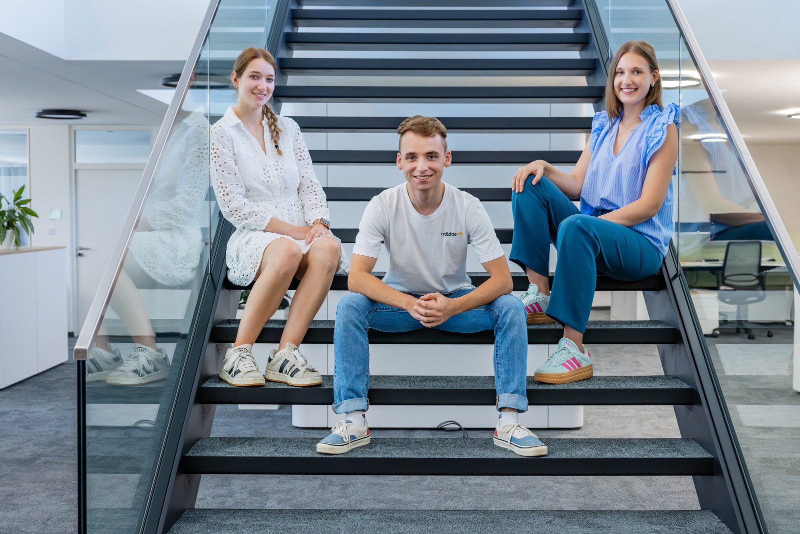Three smiling young adults sit on modern stairs in an office, looking at the camera.